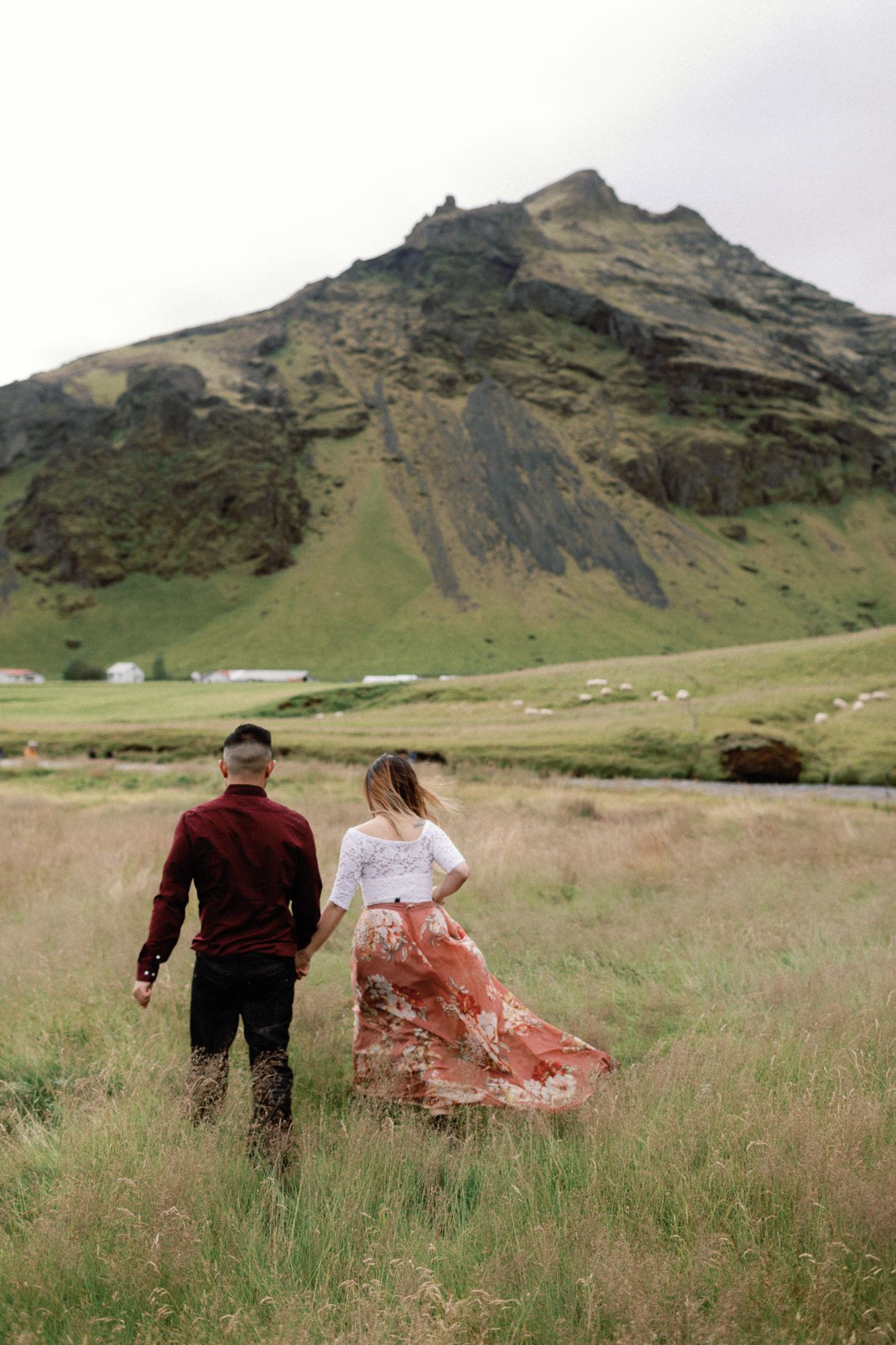 Couple walking hand in hand through tall grass fields with a dramatic green mountain in Iceland.
