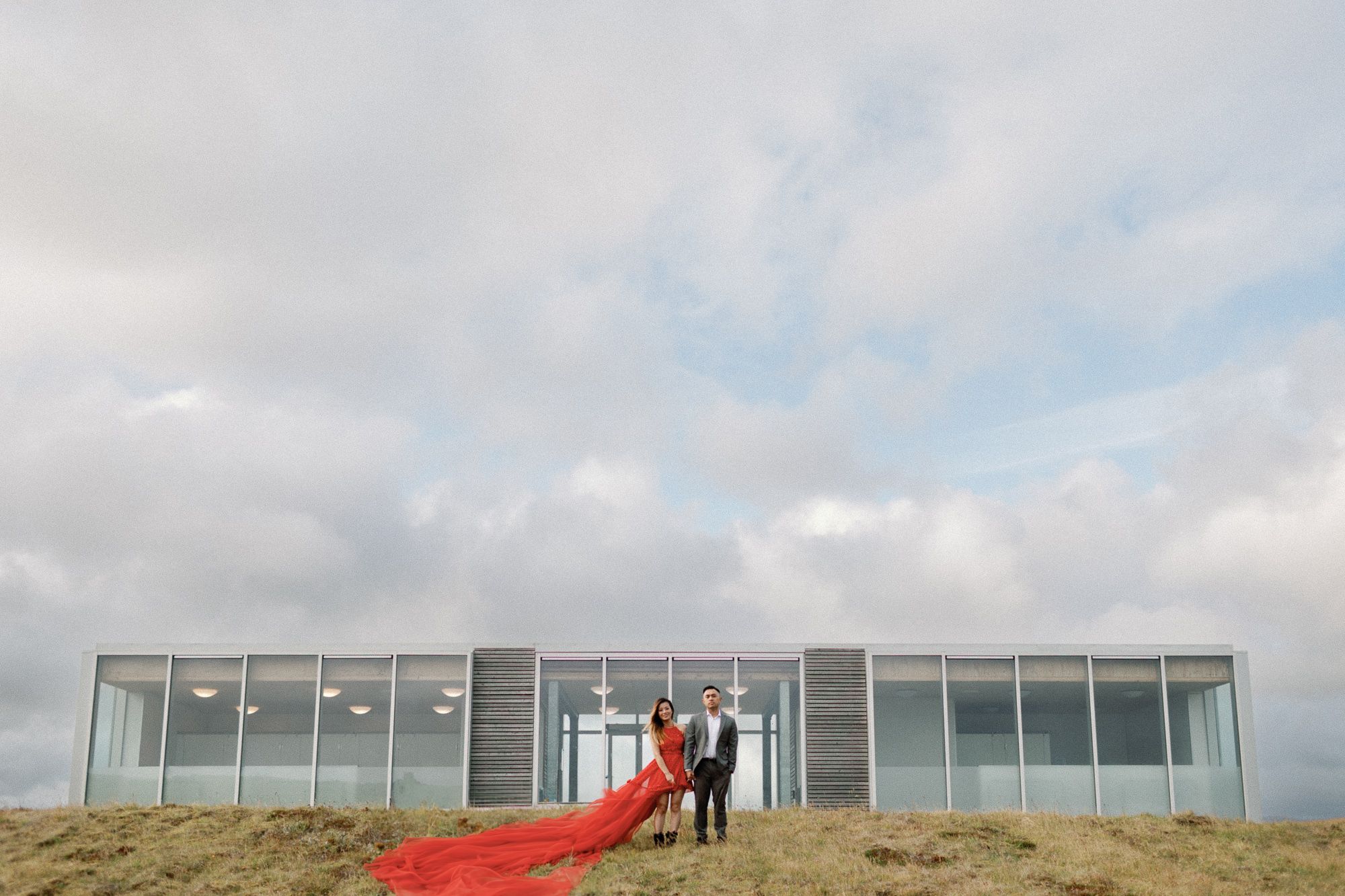 Couple standing on a grassy hill in front of a modern glass building with a dramatic red dress train flowing.