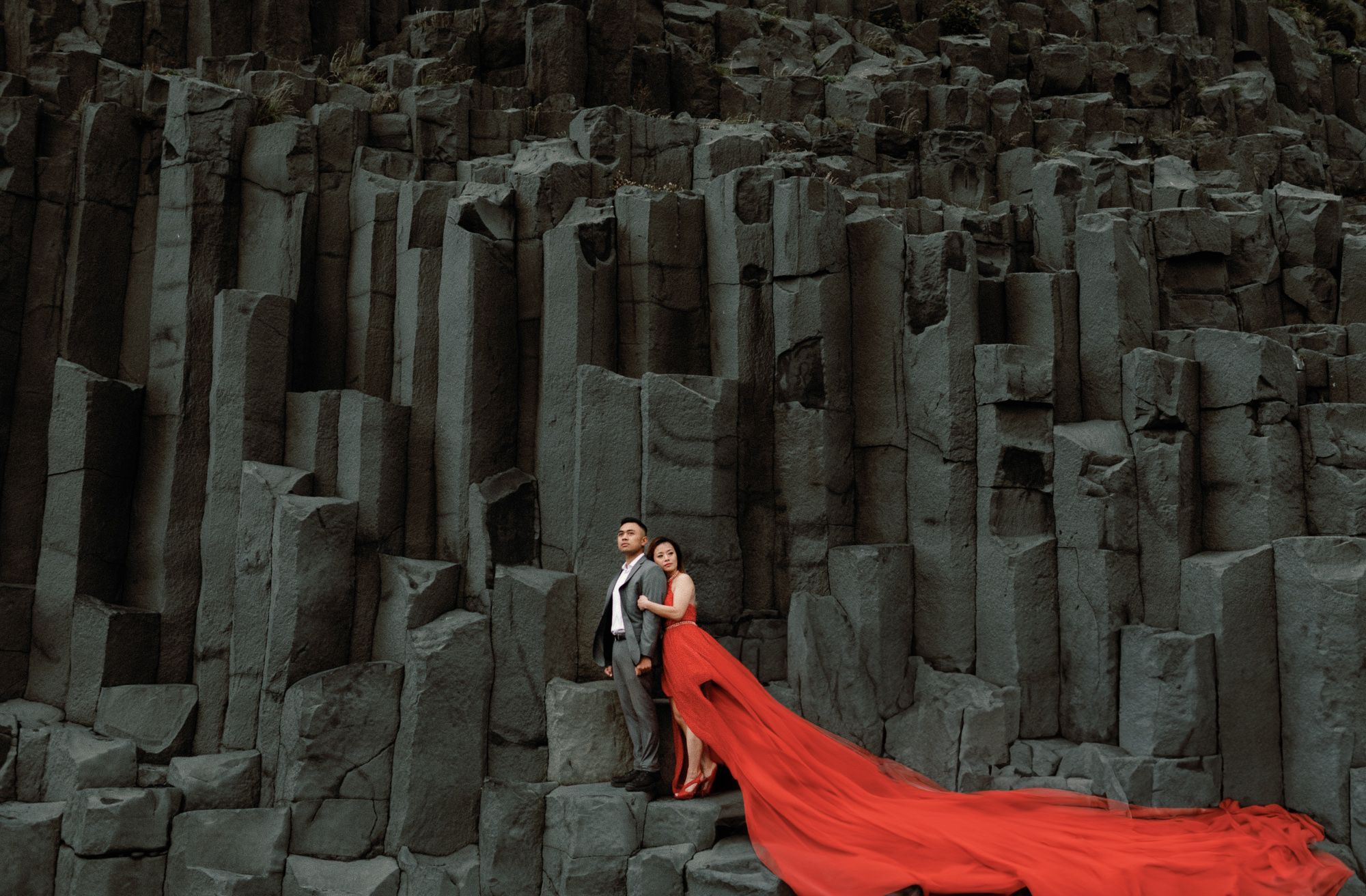 Couple embracing in a red flowing gown against dramatic dark basalt column formations at Reynisfjara beach Iceland.