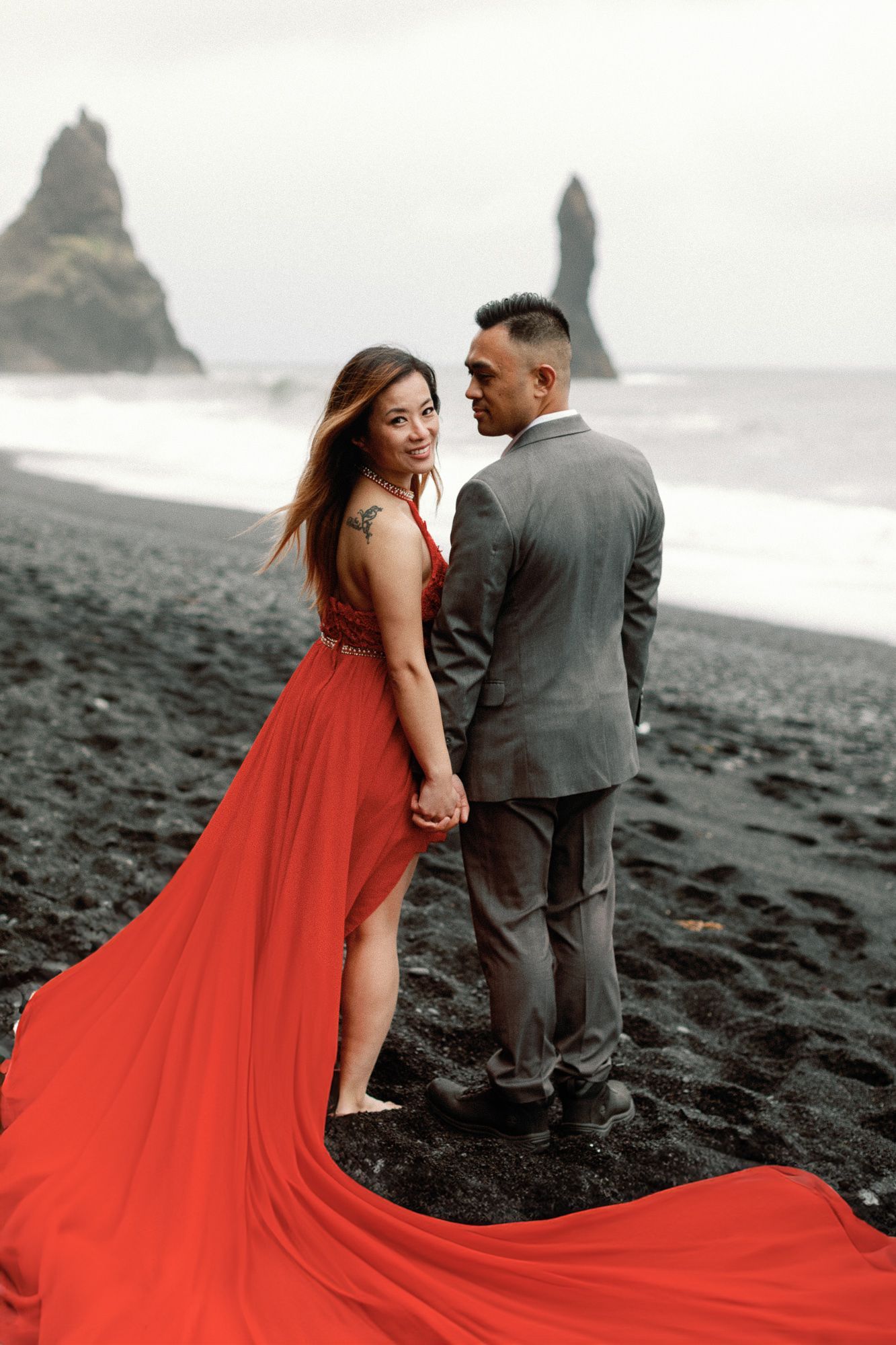 Couple on black sand beach at Reynisfjara Iceland, woman in red gown with flowing train, sea stacks in background.