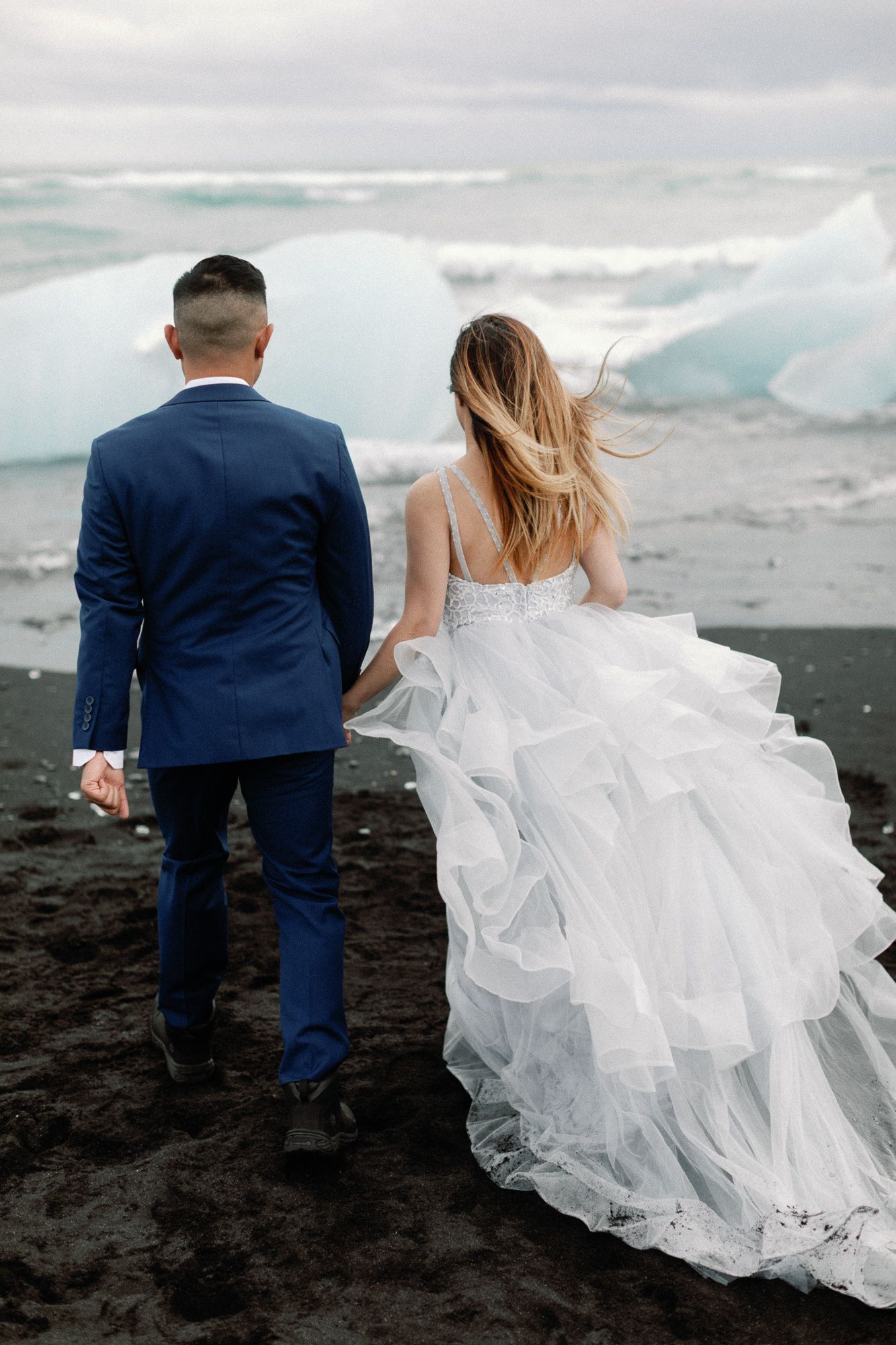 Bride and groom walking hand in hand on a black sand beach with glaciers in Iceland.