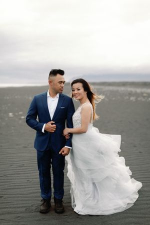 Bride in white gown and groom in blue suit standing on a vast black sand beach in Iceland.