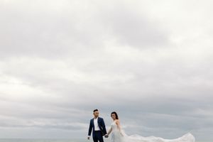 Bride and groom holding hands under a dramatic cloudy sky with the wedding dress veil flowing in the wind.
