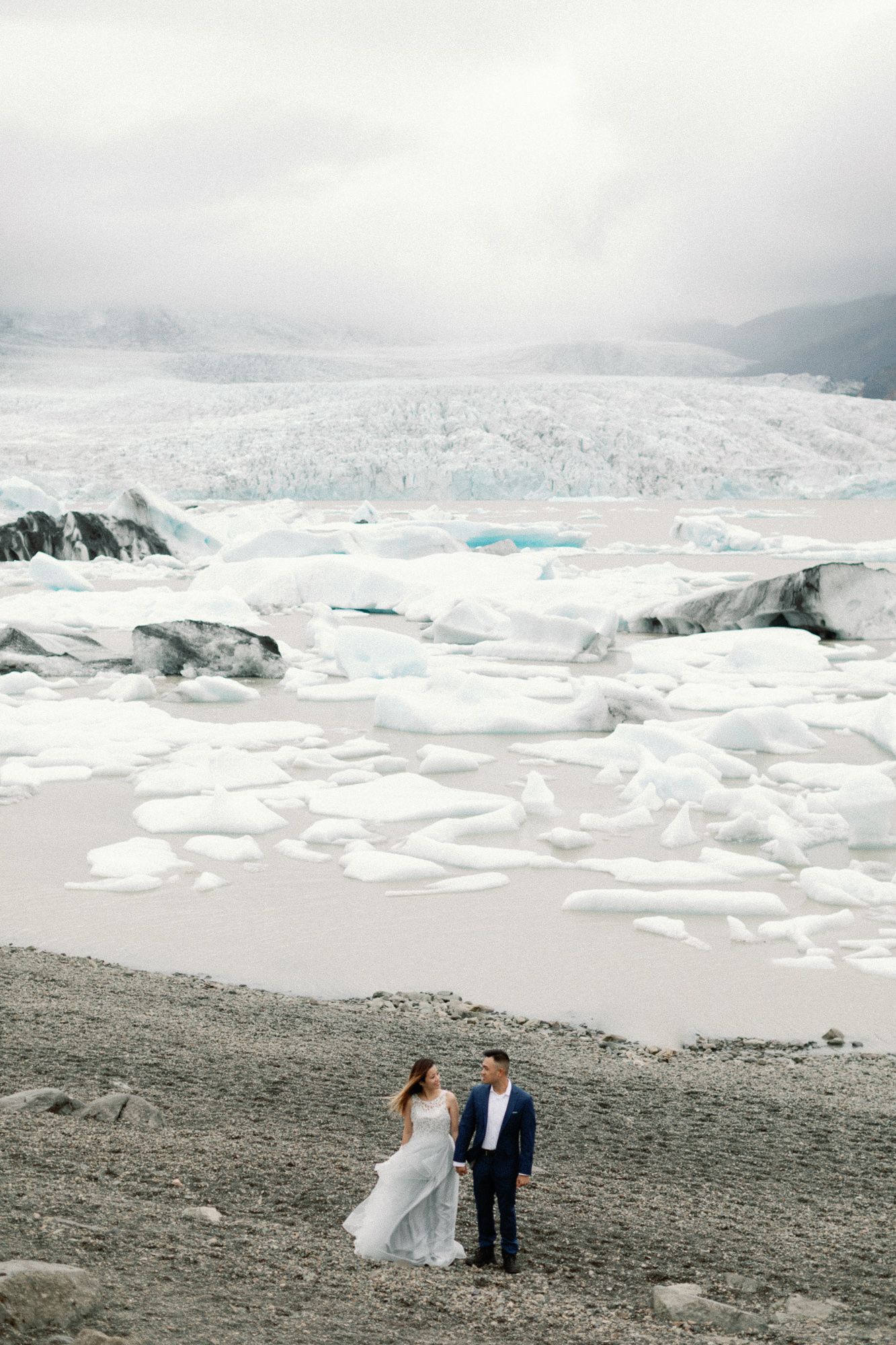 Wedding couple holding hands on a volcanic beach with a stunning glacial lagoon filled with icebergs behind them.