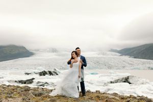 Bride and groom posing on rocky terrain with a vast glacier and mountains in the misty background in Iceland.