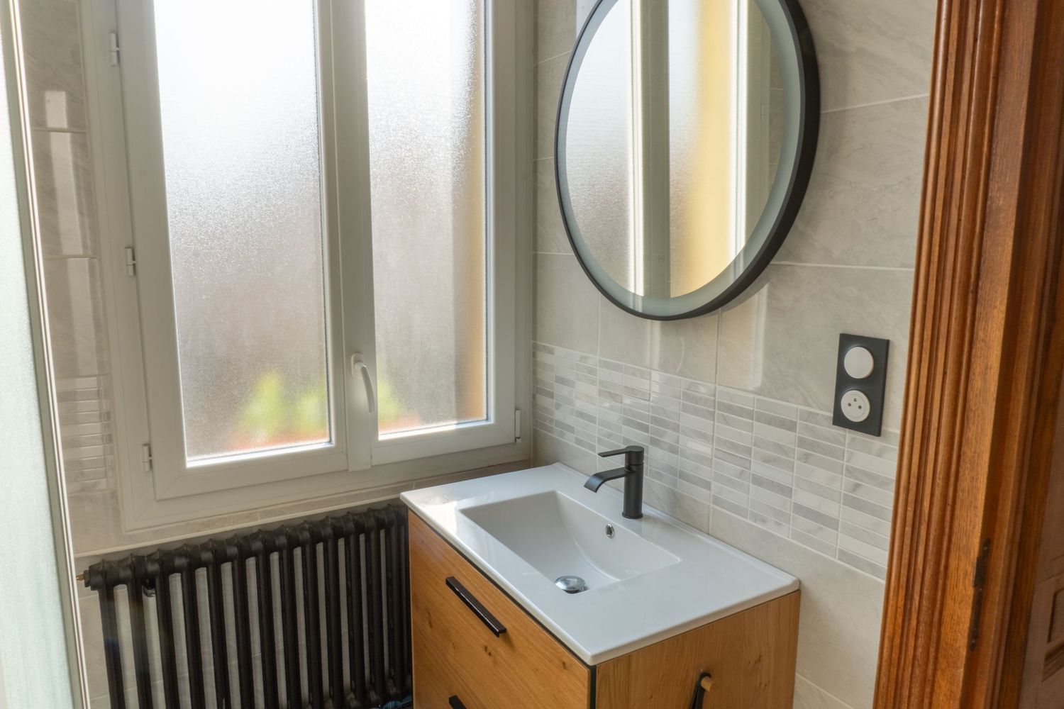 Modern bathroom vanity with wooden cabinet, white sink, round mirror, and frosted window in bright interior space.