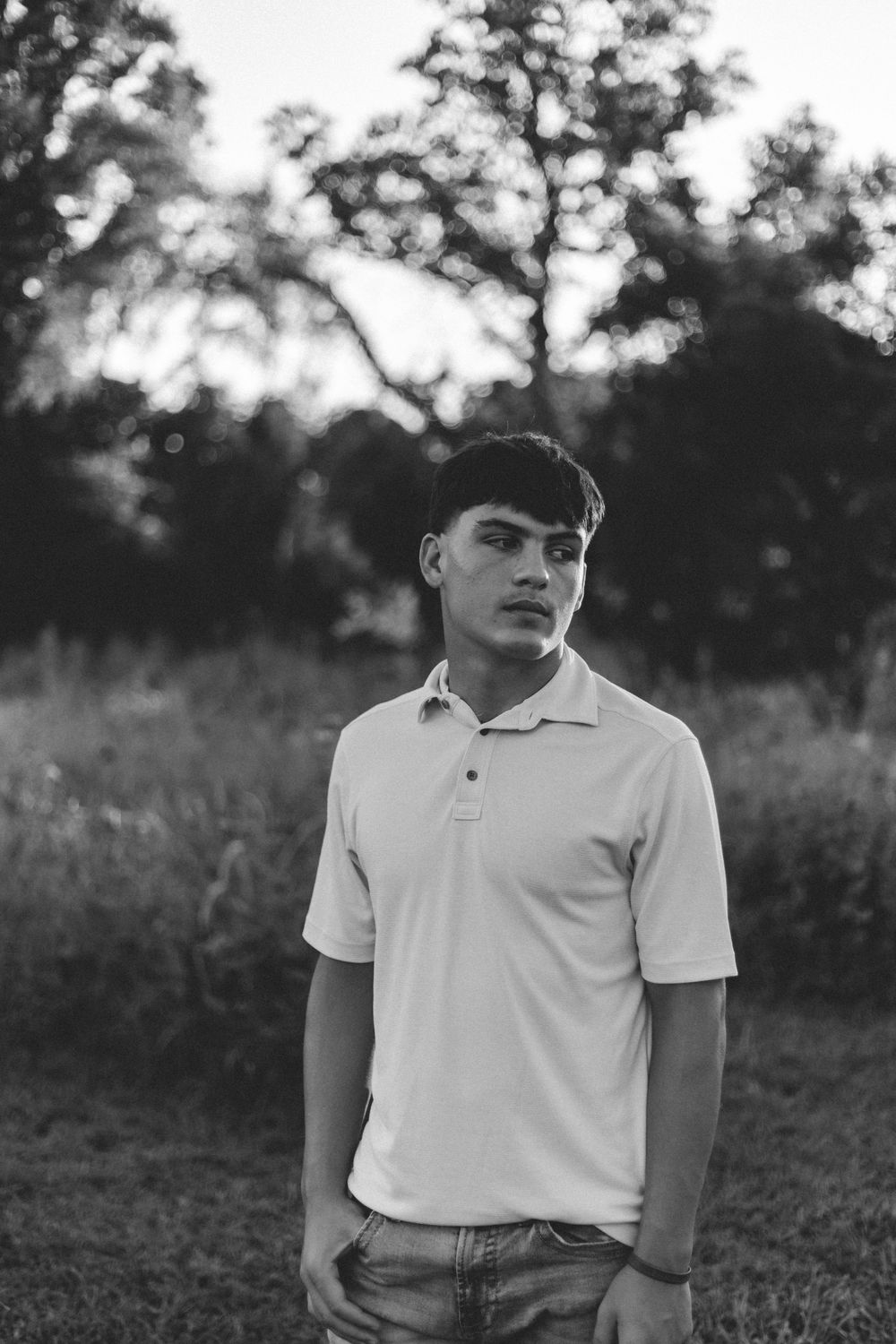 Black and white portrait of young person in polo shirt standing outdoors with trees in background at sunset.
