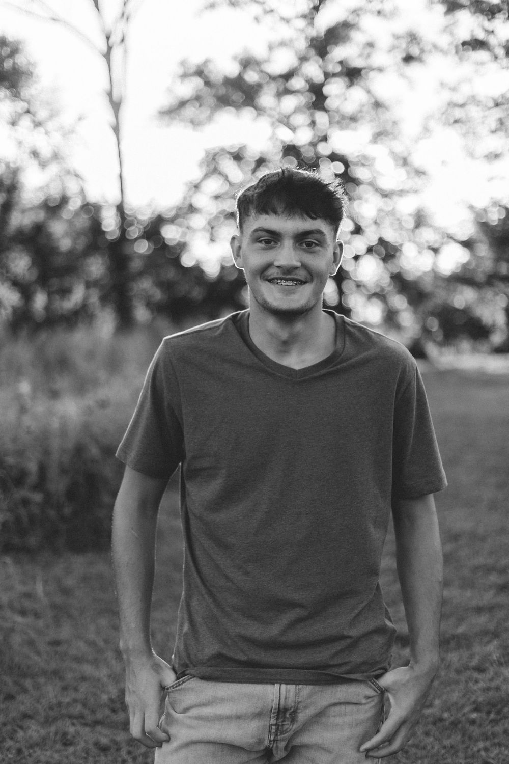 Black and white photo of casually dressed individual smiling outdoors with bokeh effect in background.