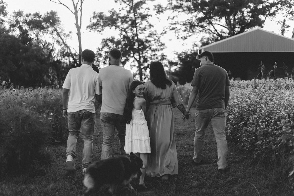 A black and white photo of a group watching a sunset together in a wildflower field near a wooded area.