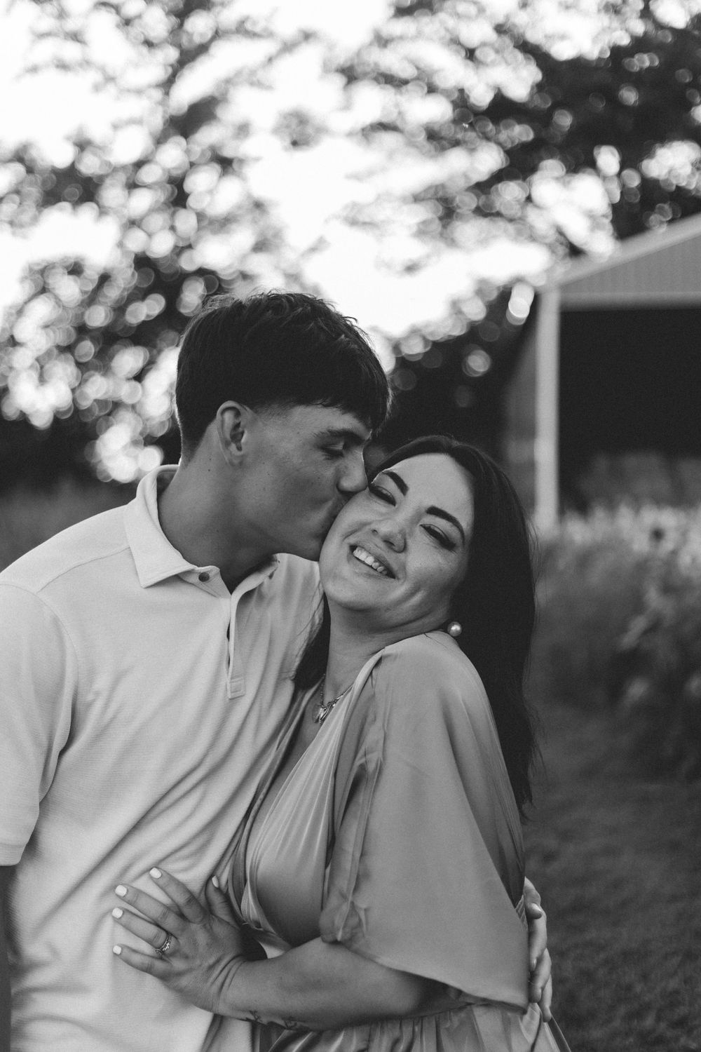 A romantic black and white portrait of a couple laughing and embracing outdoors with soft bokeh lighting in the background.