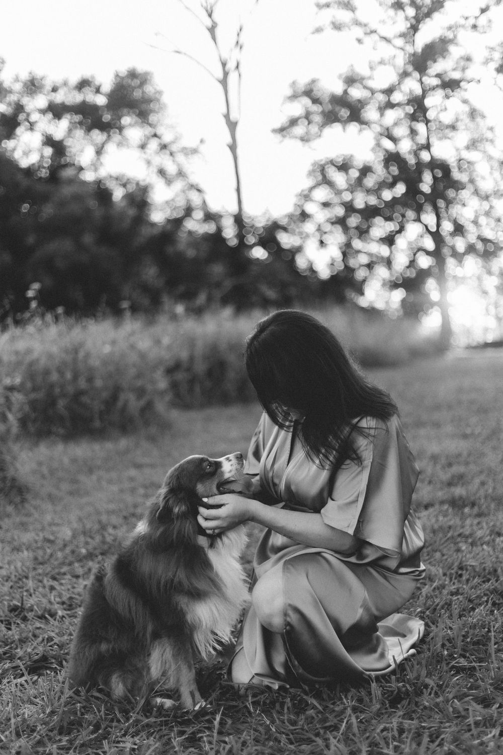 A person in a flowing dress sits in a grassy field petting their Australian Shepherd dog at sunset.
