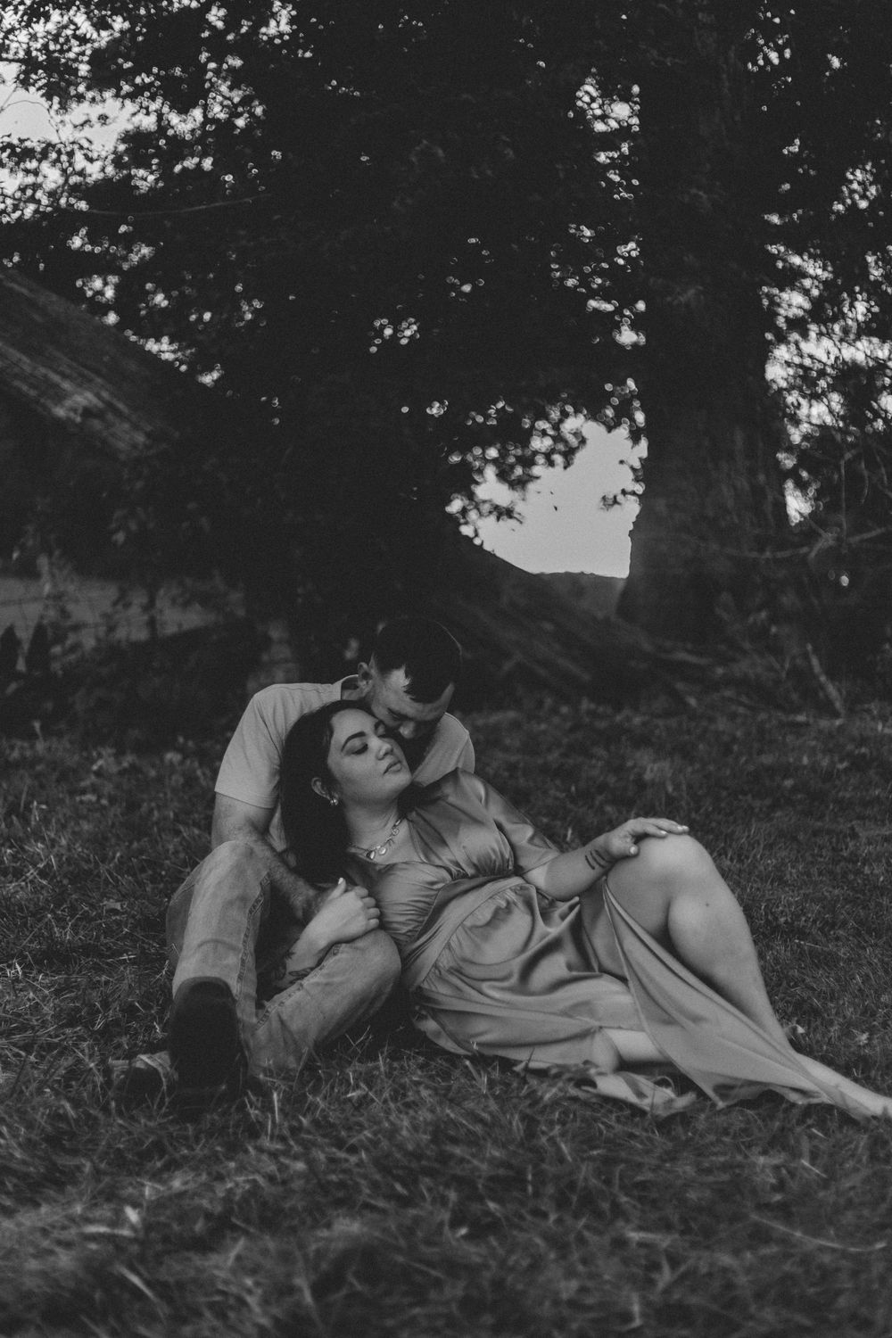 A romantic black and white photograph of a couple resting together beneath a tree in nature.
