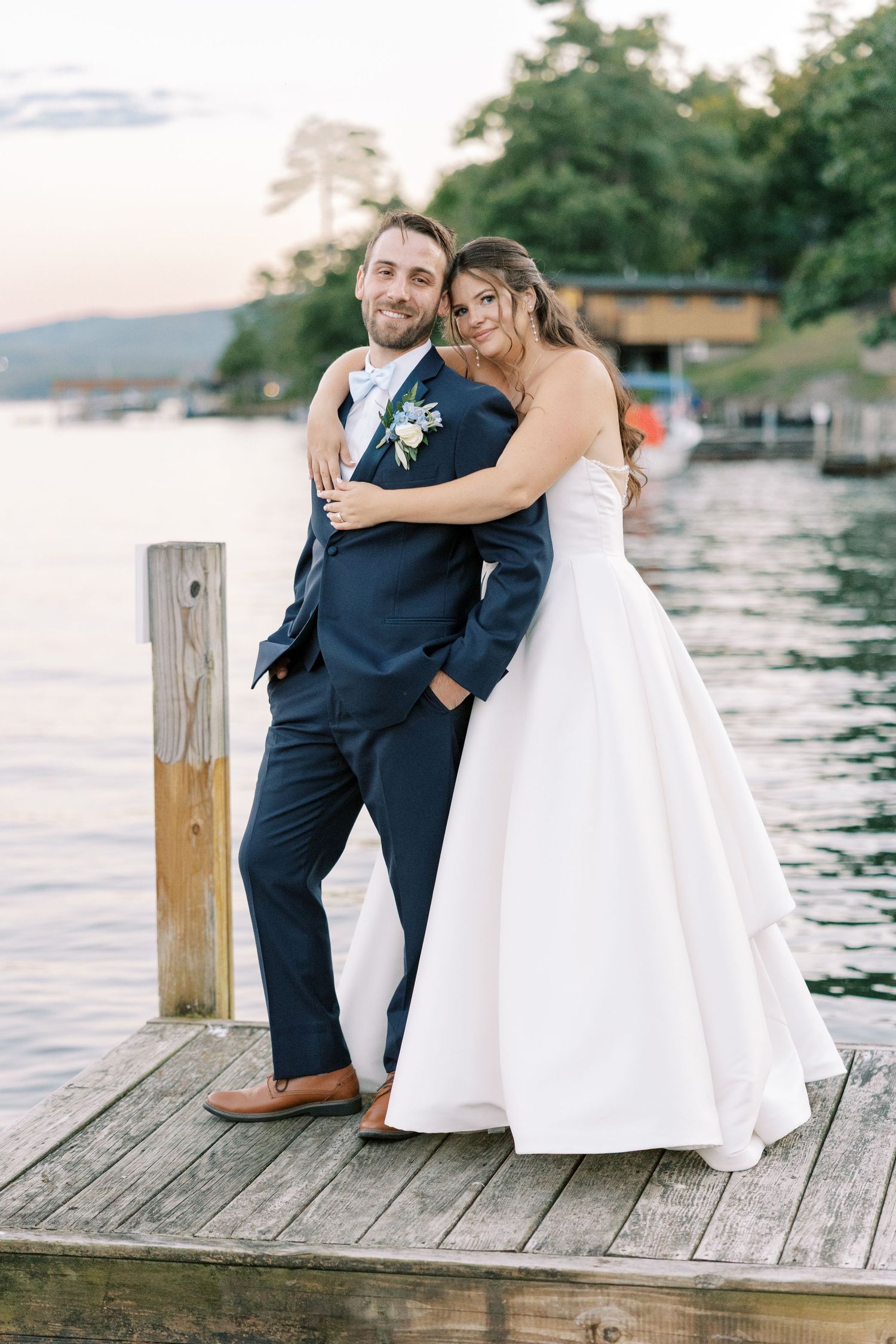 Couple in wedding attire sitting together on wooden dock by tranquil lake at sunset.
