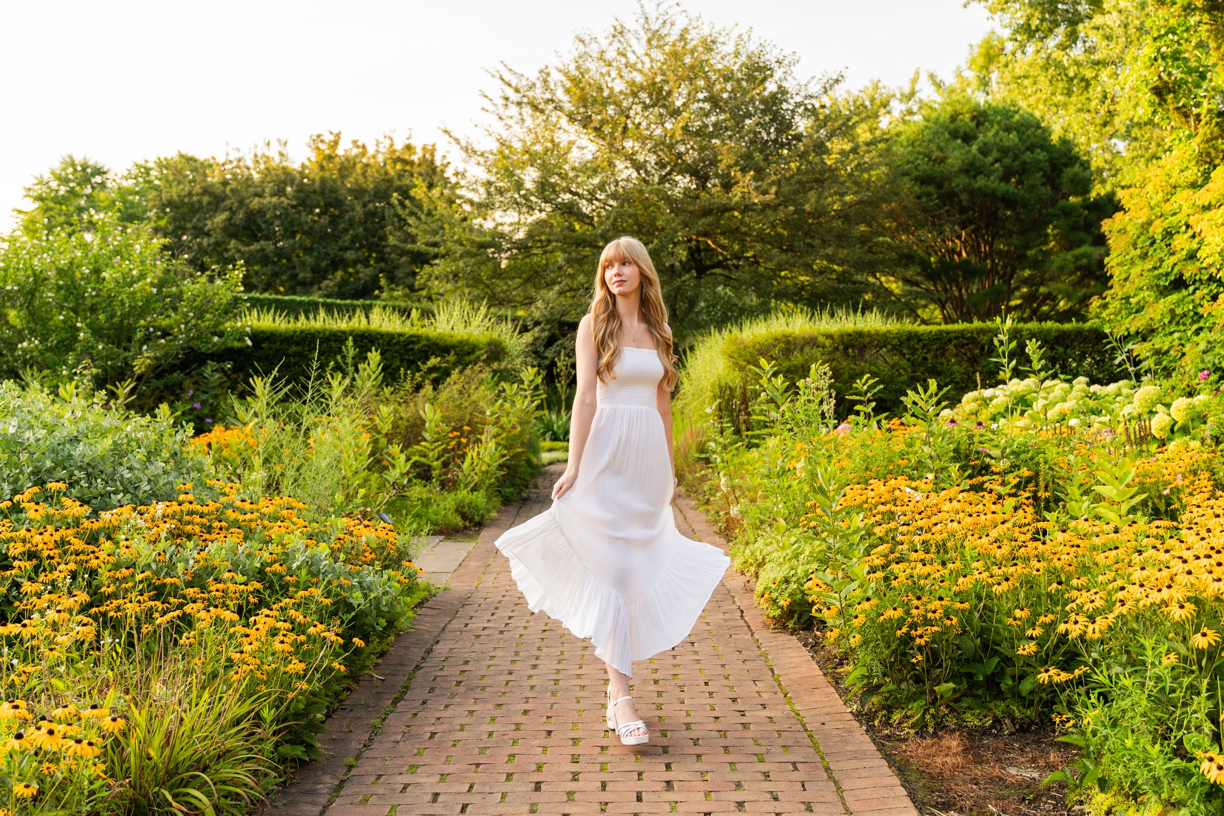 A figure in a flowing white dress twirls on a garden path surrounded by vibrant yellow flowers and greenery at sunset.