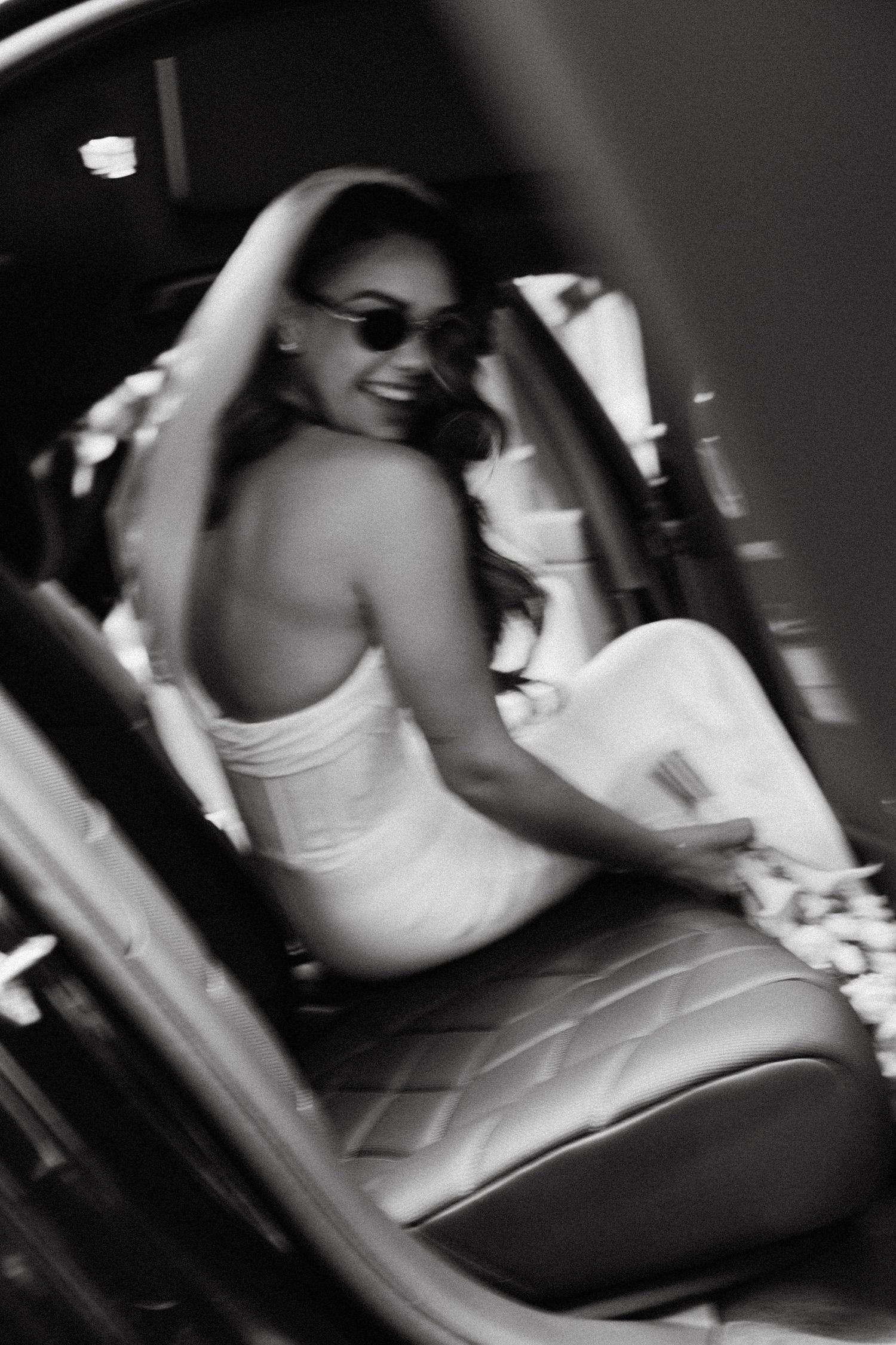 Black and white photo of someone wearing sunglasses looking back over their shoulder while sitting in a car seat.