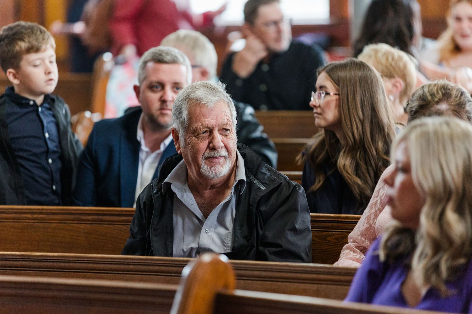 People sit in wooden pews inside a church during a service or gathering.