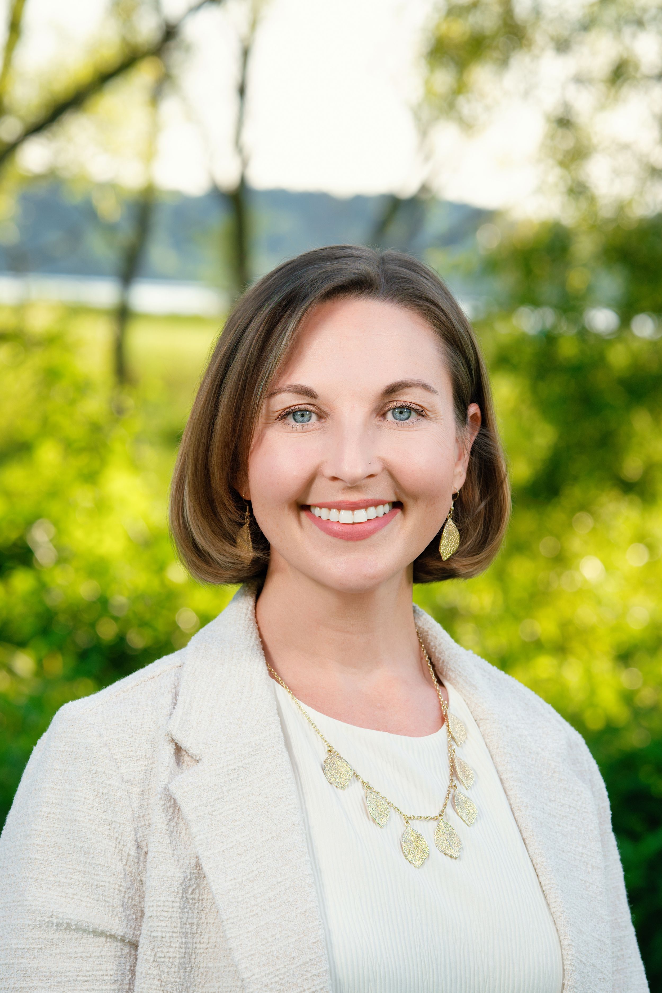 Professional portrait in natural lighting featuring a warm smile and white blazer against a blurred green outdoor background.