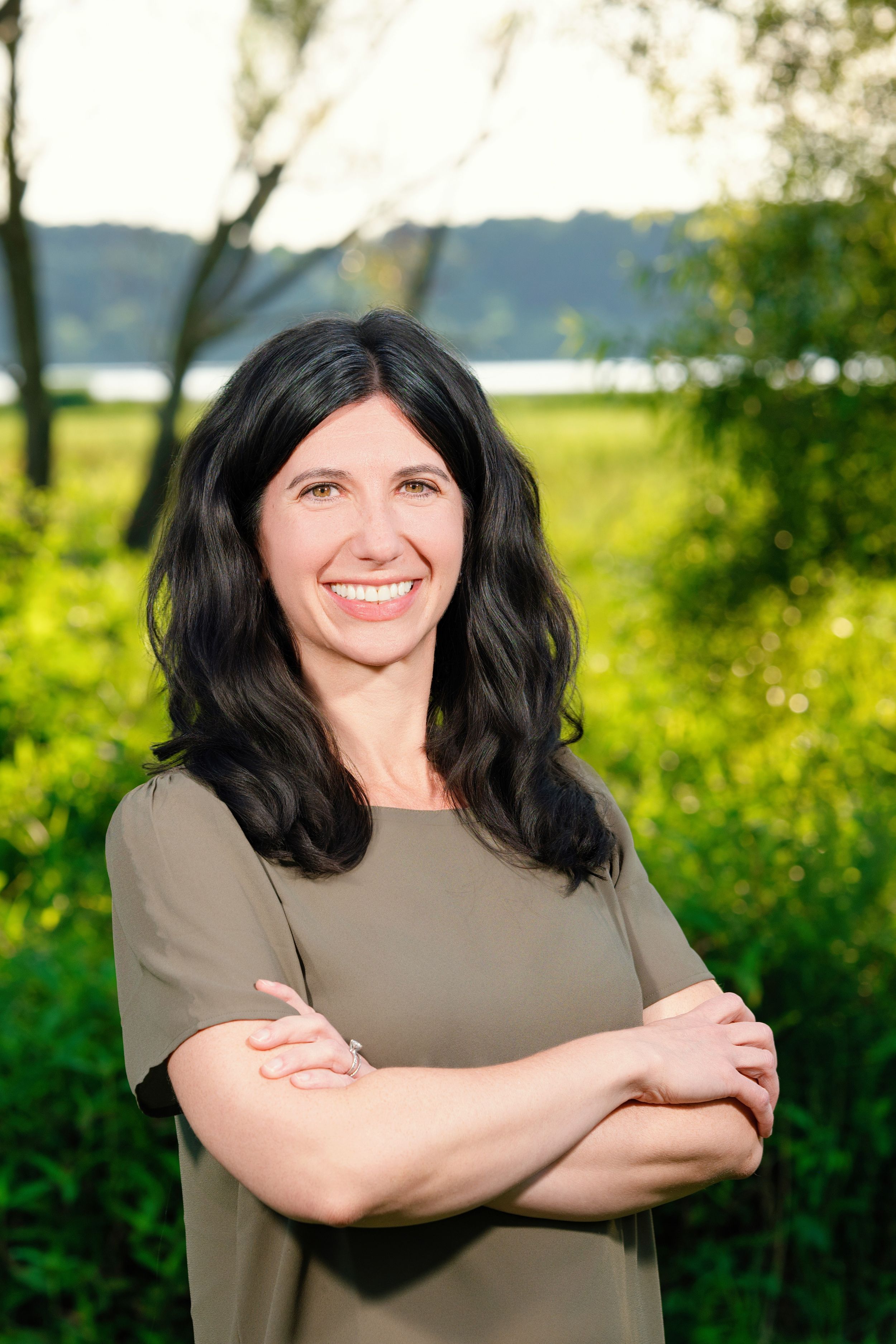 A woman with dark hair smiles while posing outdoors against a natural green background on a sunny day.