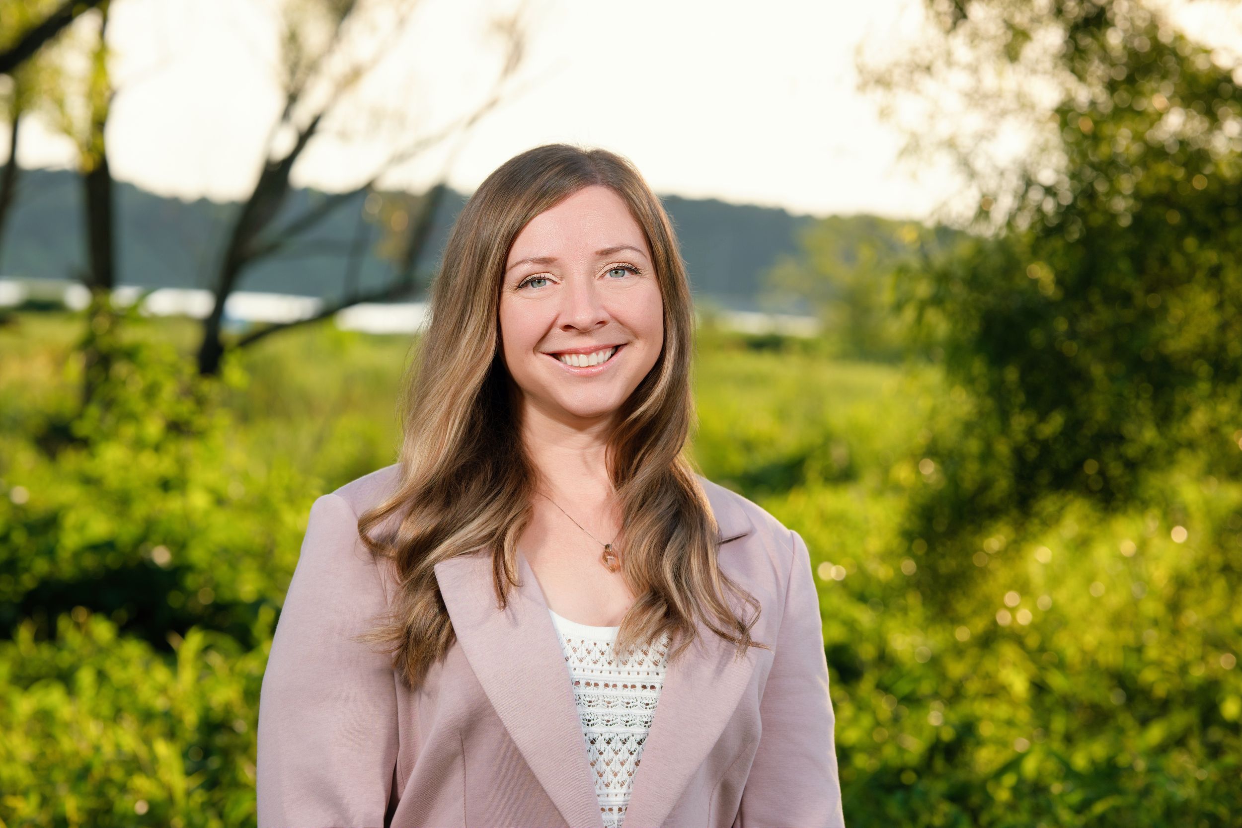 A person wearing a gray blazer smiles while posing outdoors in a natural setting during golden hour.