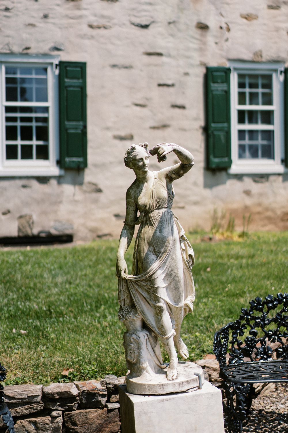A classical stone garden statue stands beside a stone wall with green shuttered windows in the background.