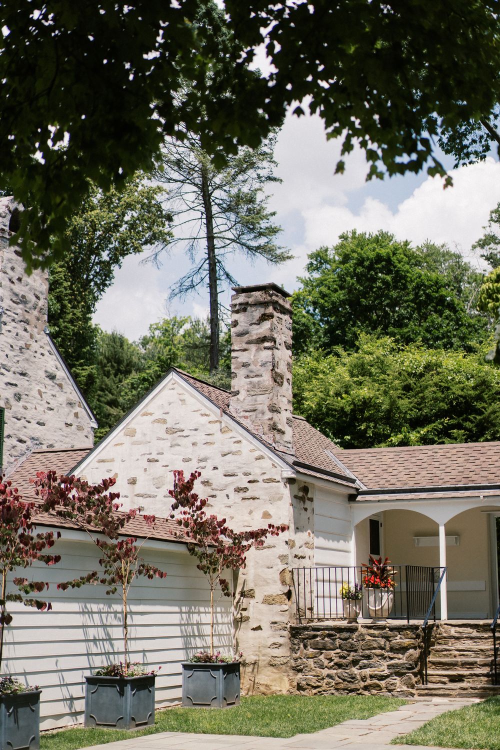 Stone cottage with white siding and brick chimneys sits nestled among trees in a garden setting.