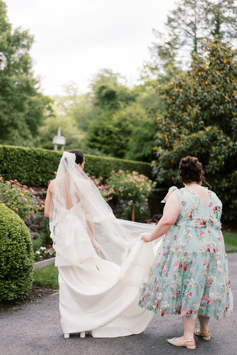 Two attendants in floral dresses help carry a flowing white wedding dress train through a garden path.