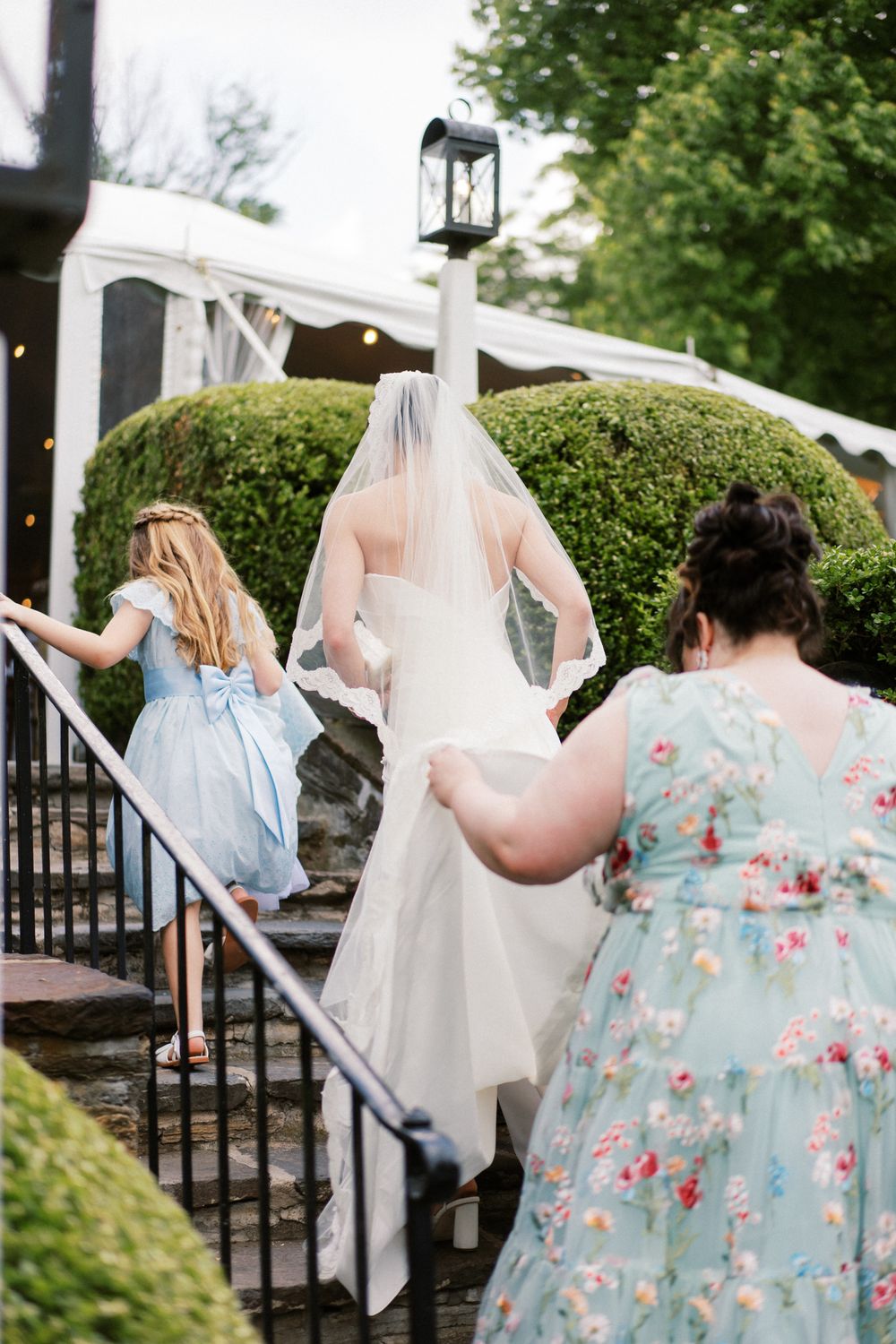 Wedding party members assist with a bridal veil on outdoor stone steps.