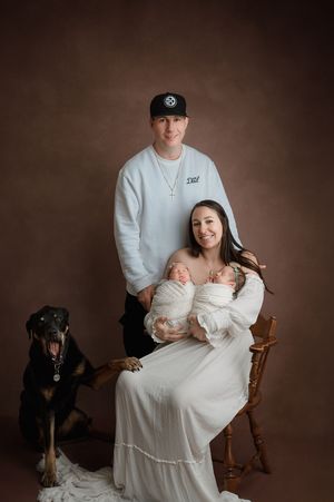 A newborn photo session featuring a baby in white with parents and black dog in a warm studio setting.