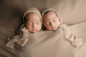 Two newborn babies wrapped in beige blankets rest peacefully side by side on neutral backdrop.