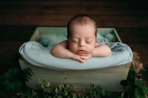Newborn baby sleeps peacefully in cream-colored blankets surrounded by green ferns and natural light.