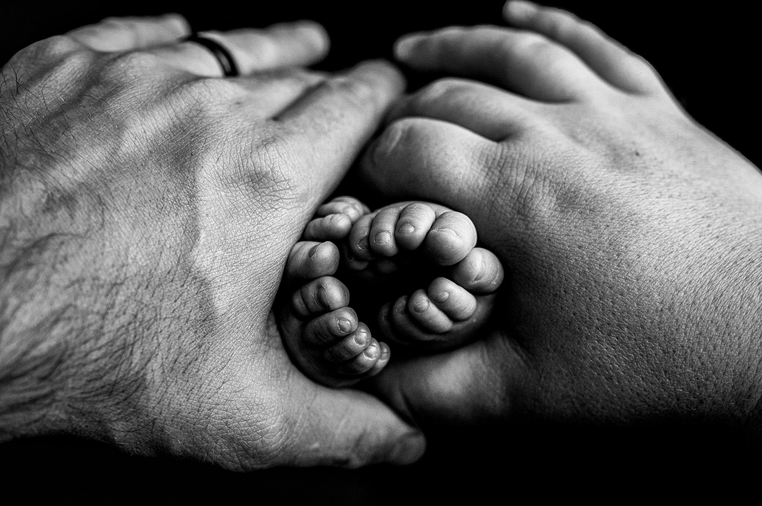 A tiny baby fist being gently held between two adult hands in black and white photography.