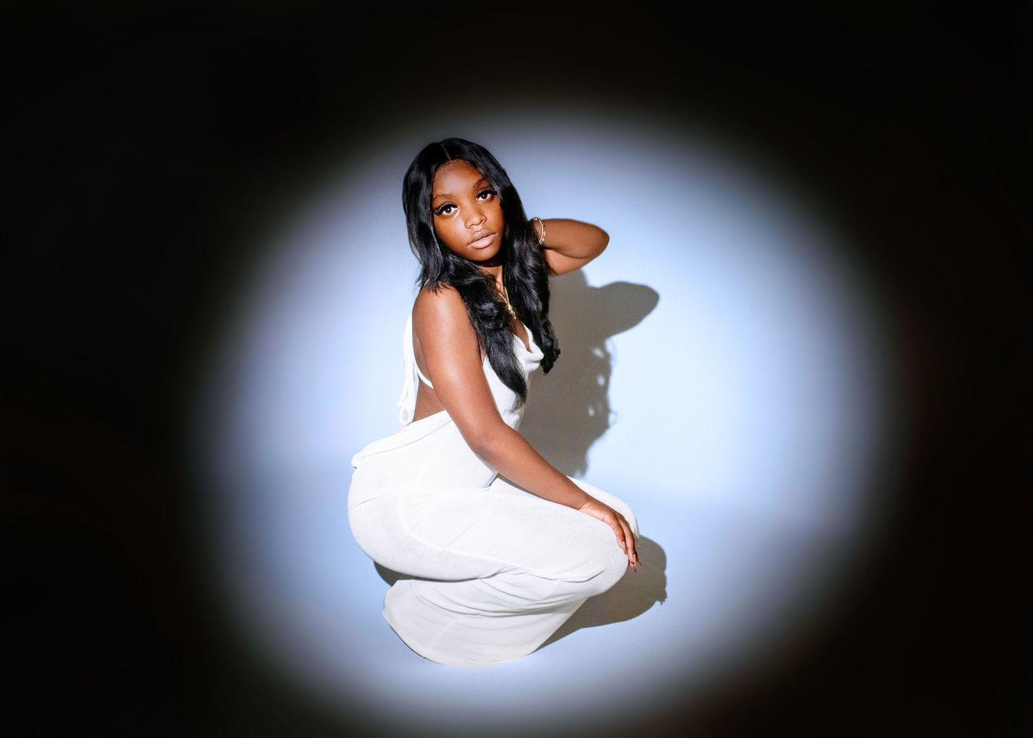 Figure in a white outfit poses seated against dark background with dramatic circular lighting effect.