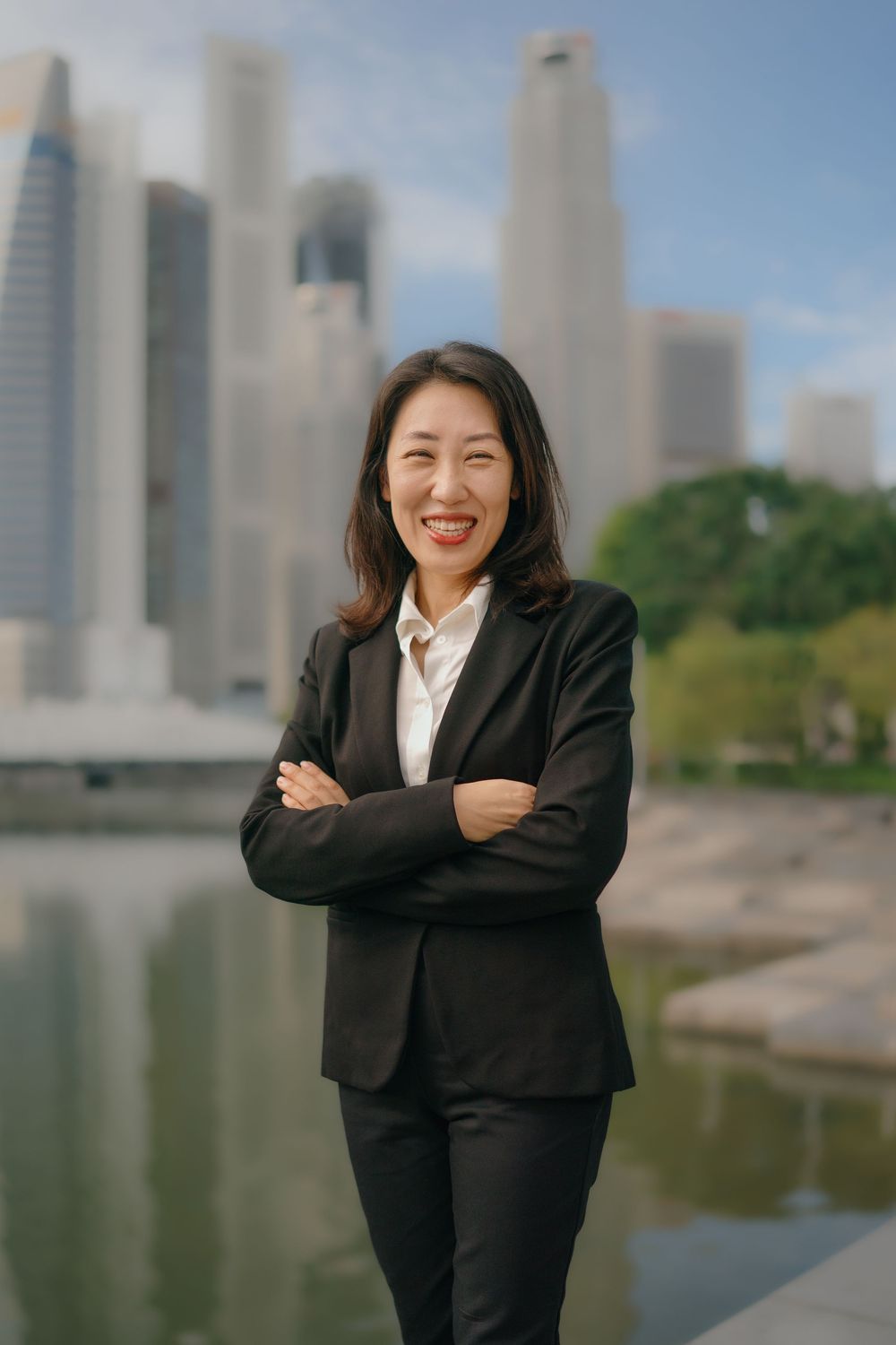 Business professional poses outdoors against modern city skyline and reflecting pool in black suit.