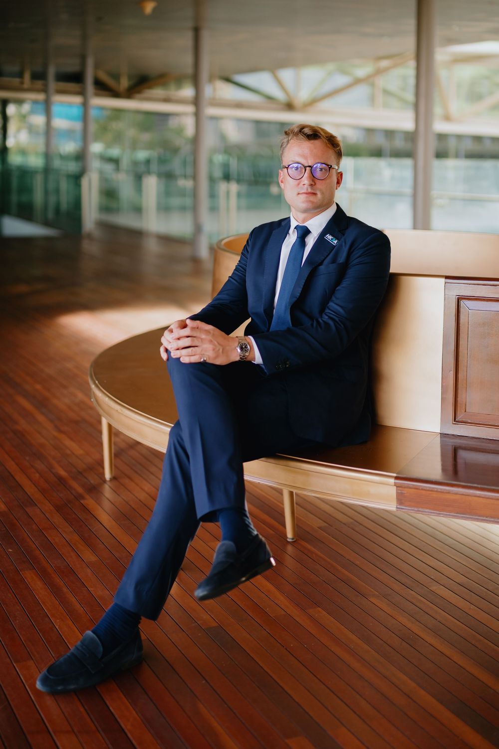 Professional in a navy suit sits on curved wooden bench while looking at smartphone in modern office setting.
