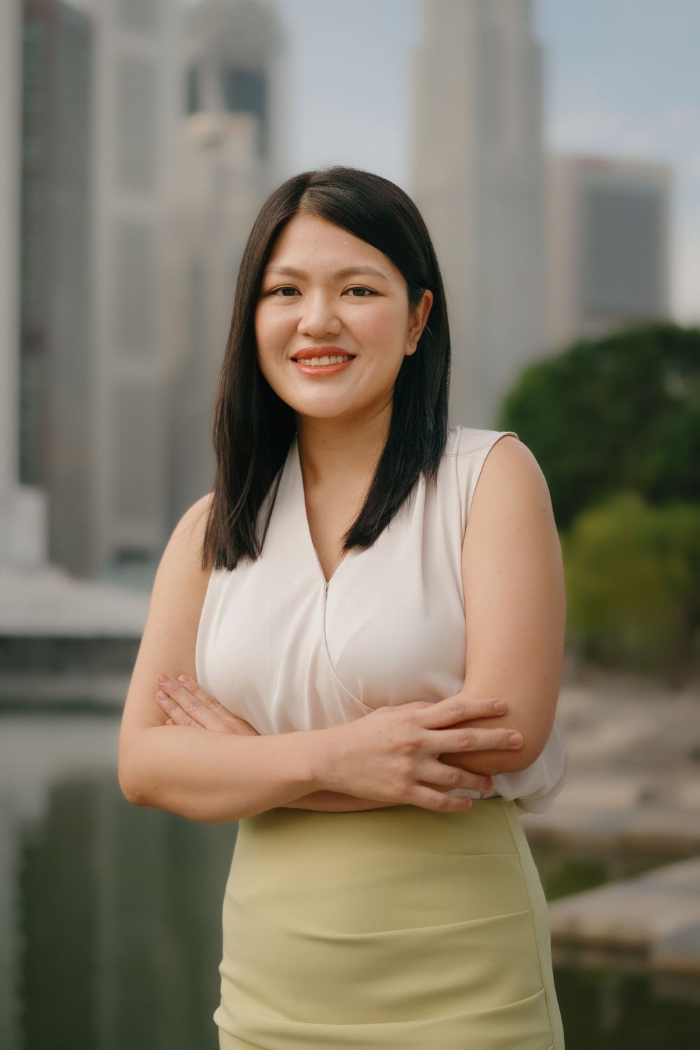 Asian woman in white sleeveless top and light skirt stands confidently with city buildings in the background.