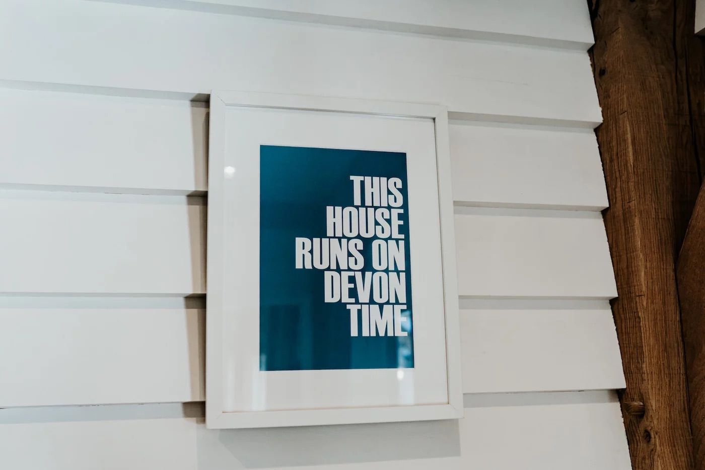 A blue and white sign mounted on white siding reads 'This House Runs on Devon Time'.