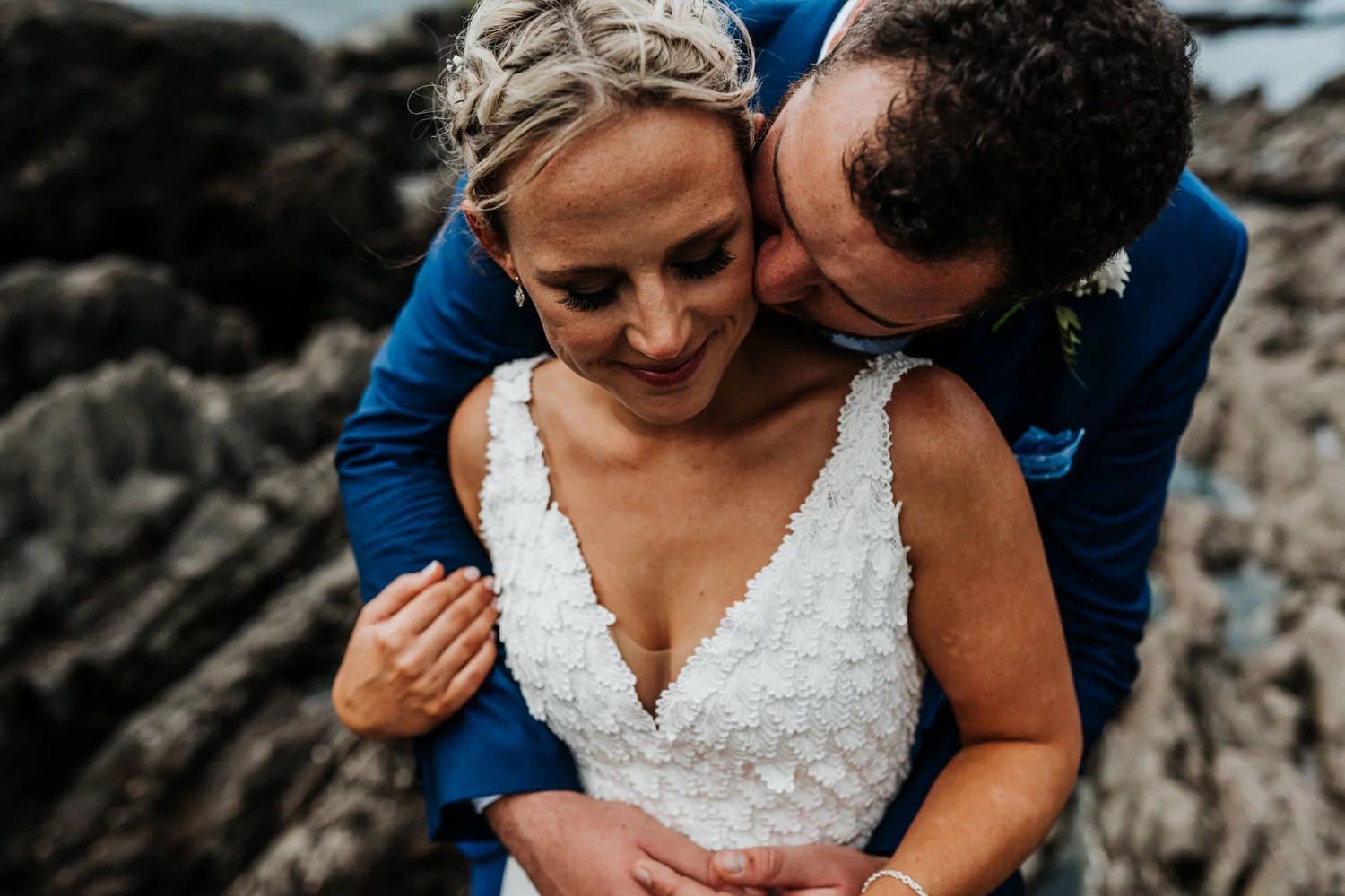 An intimate romantic moment captured between a couple in formal attire against a rocky beachside backdrop.