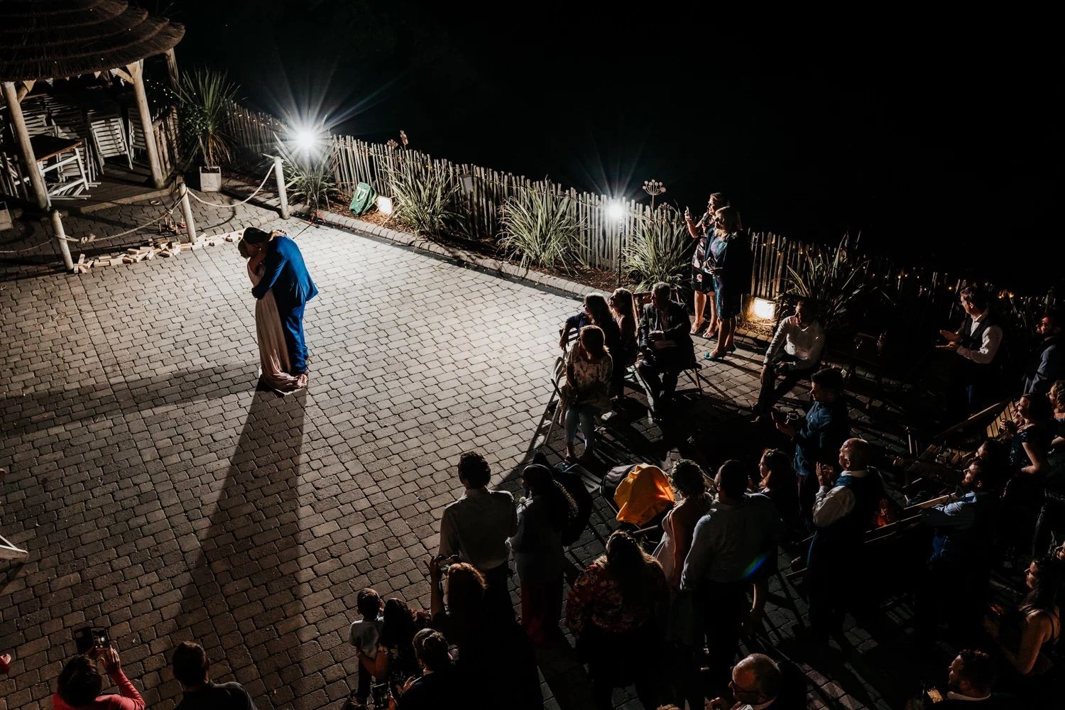 A crowd gathers at night around a lit outdoor venue with cobblestone flooring and ambient lighting.