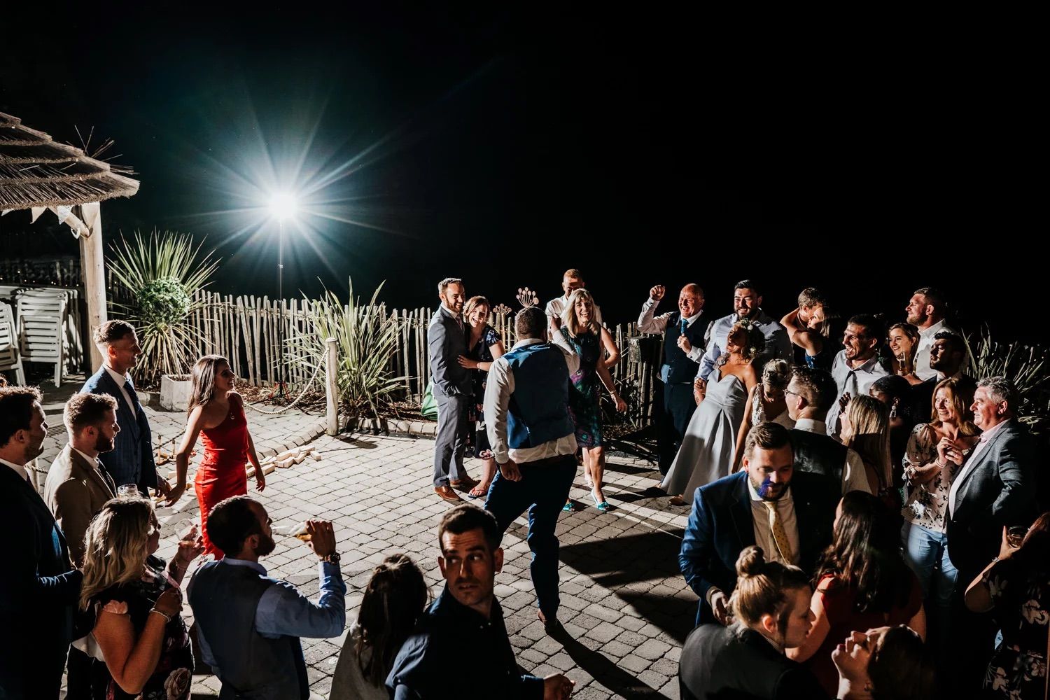 Wedding guests celebrate outdoors with bright sunburst overhead during daytime celebration.
