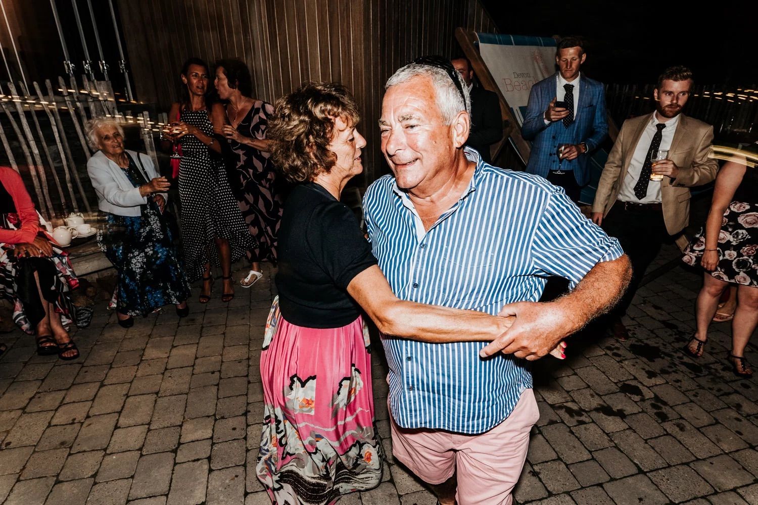 Couples dancing together at a lively evening celebration with festive lighting and decorations in the background.