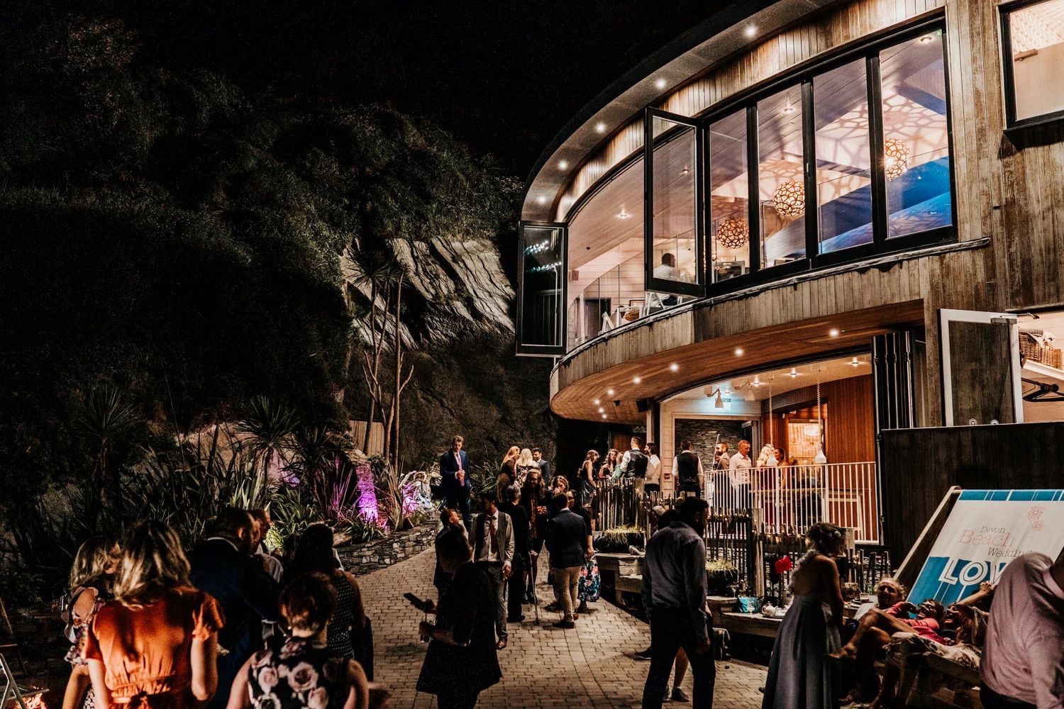 Nighttime scene outside a curved modern building with warm lighting and people gathered around the entrance.