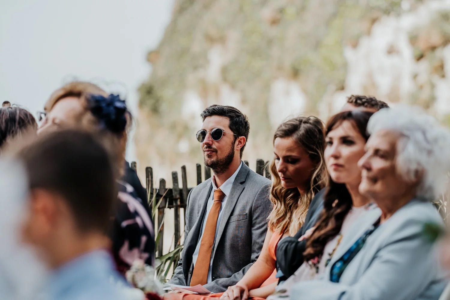 Wedding guests seated outdoors watching ceremony against mountain backdrop wearing sunglasses.