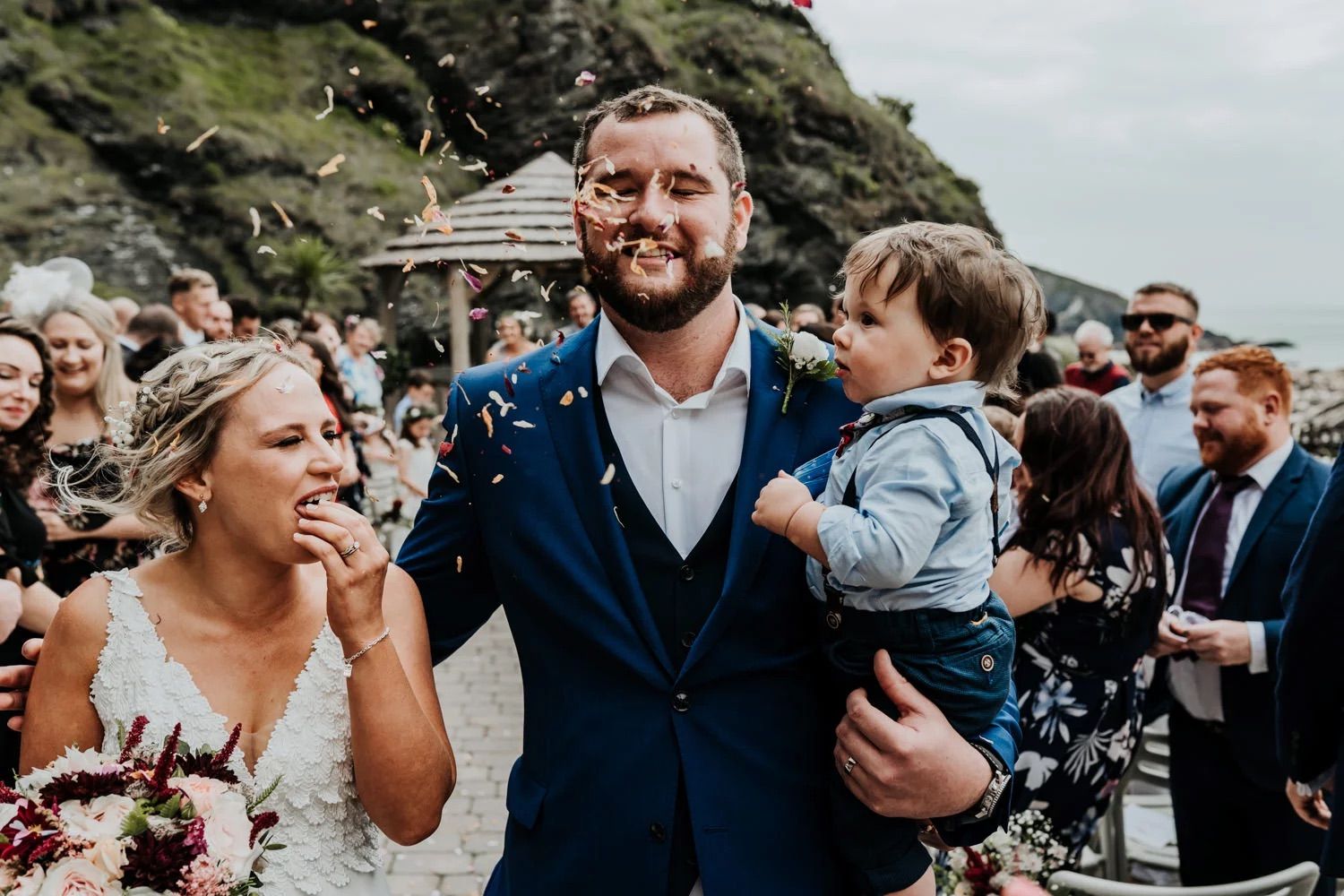 Happy wedding celebration on a rocky coastal cliffside with guests gathered around the couple.