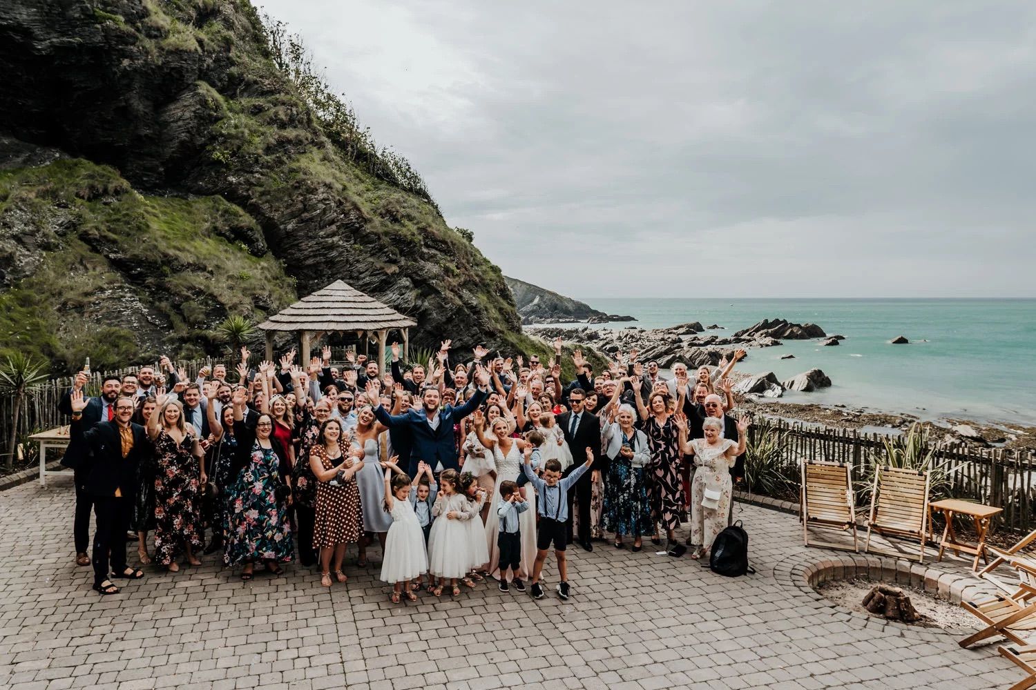 Large wedding party and guests gather for a group photo on a scenic coastal cliff overlook.