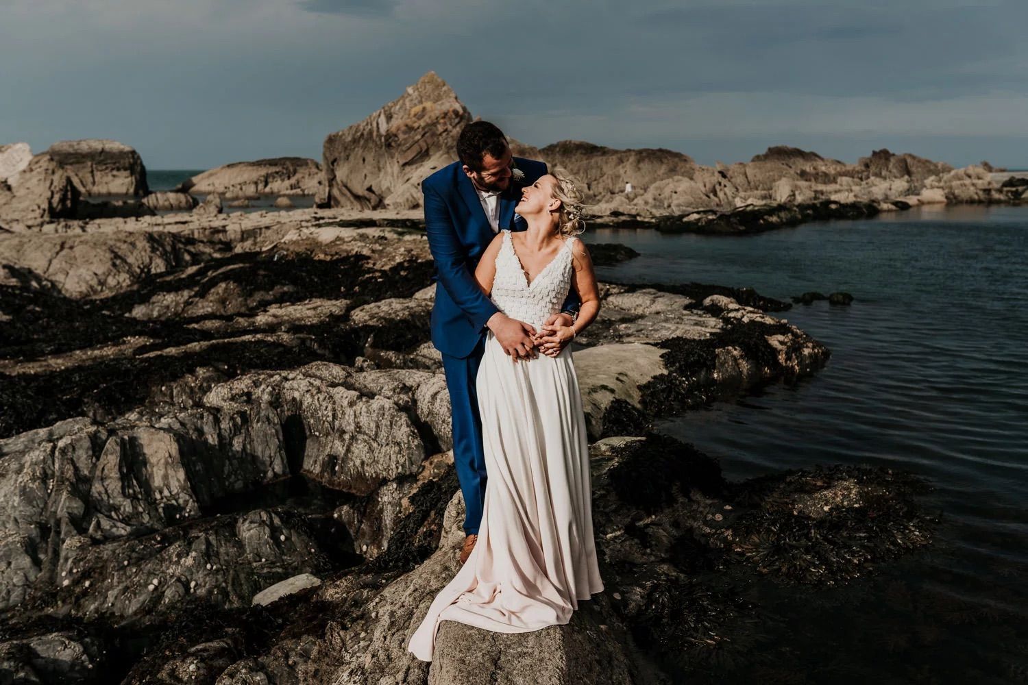 A couple embraces on rocky coastal cliffs overlooking a dramatic seascape at sunset.