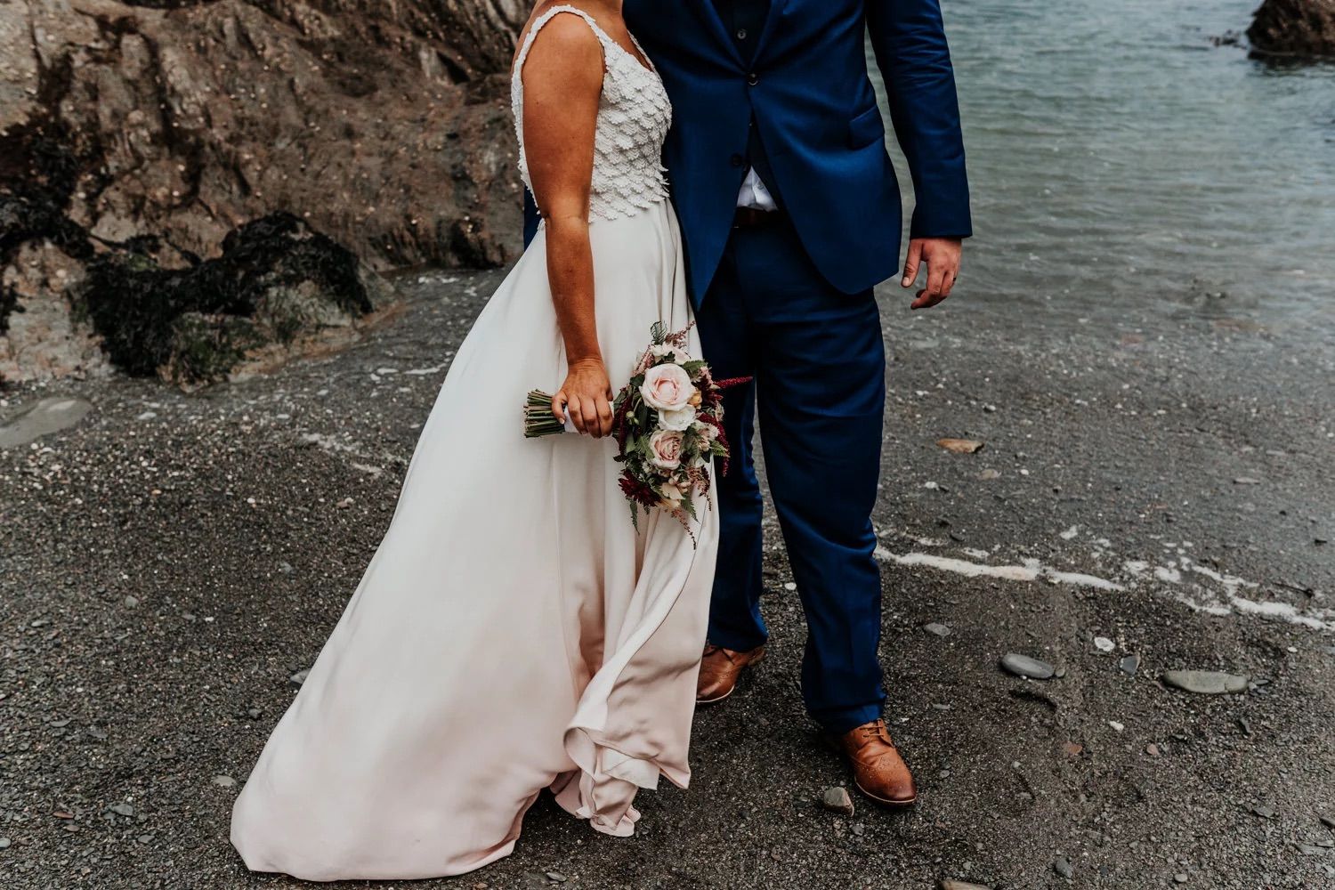 A bride and groom walk along a rocky beach coastline during their intimate wedding celebration.