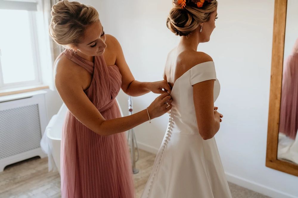 Bridesmaid in pink adjusts wedding dress details in a bright white room.