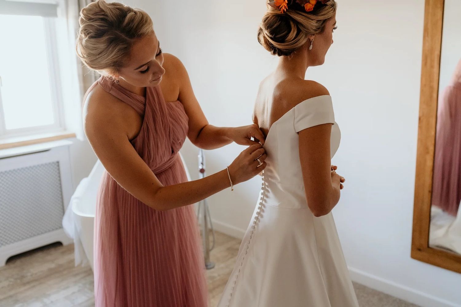 Bridesmaid in pink adjusts wedding dress details in a bright white room.
