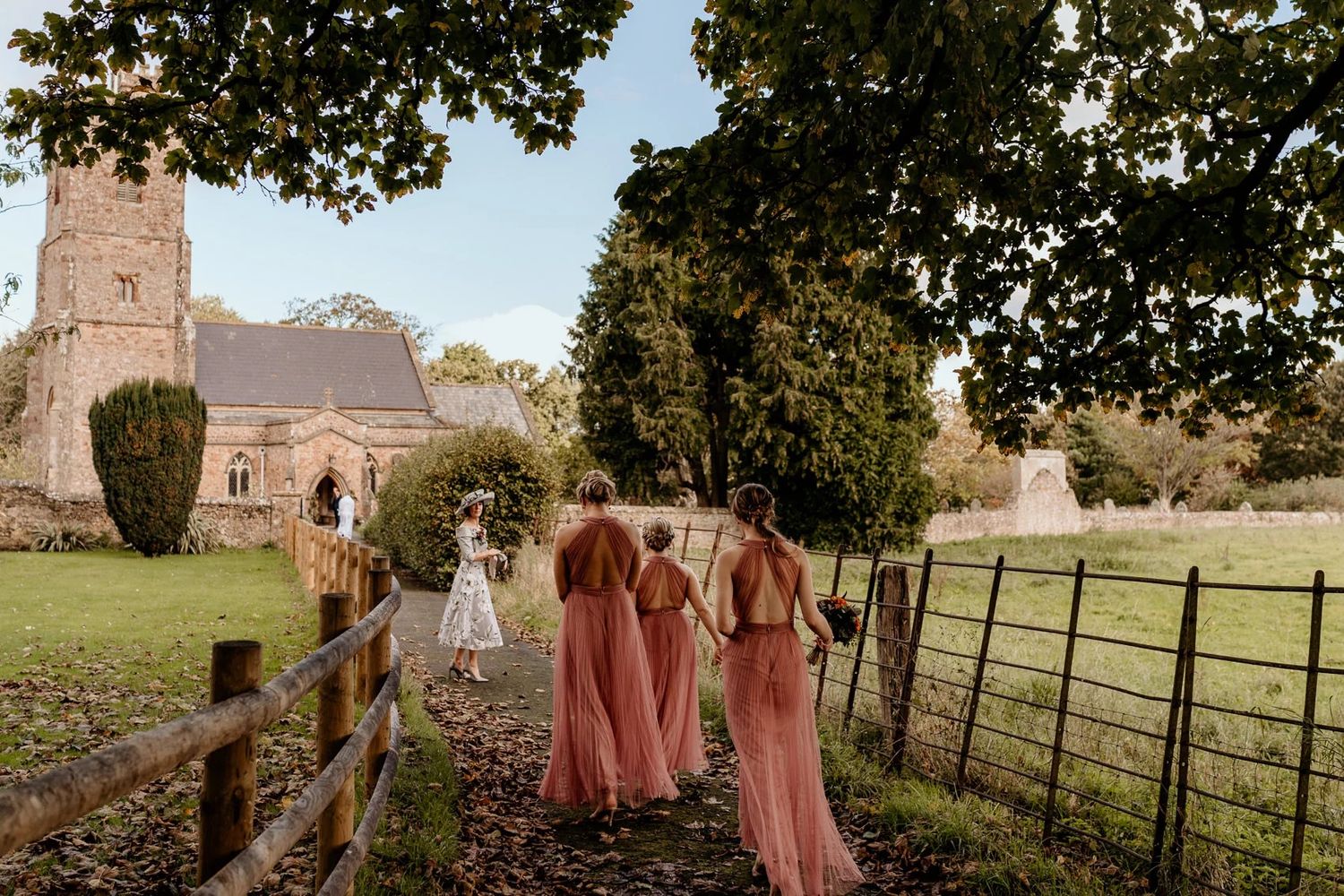 Wedding party members in pink dresses walk along a path leading to a stone church on a sunny day.
