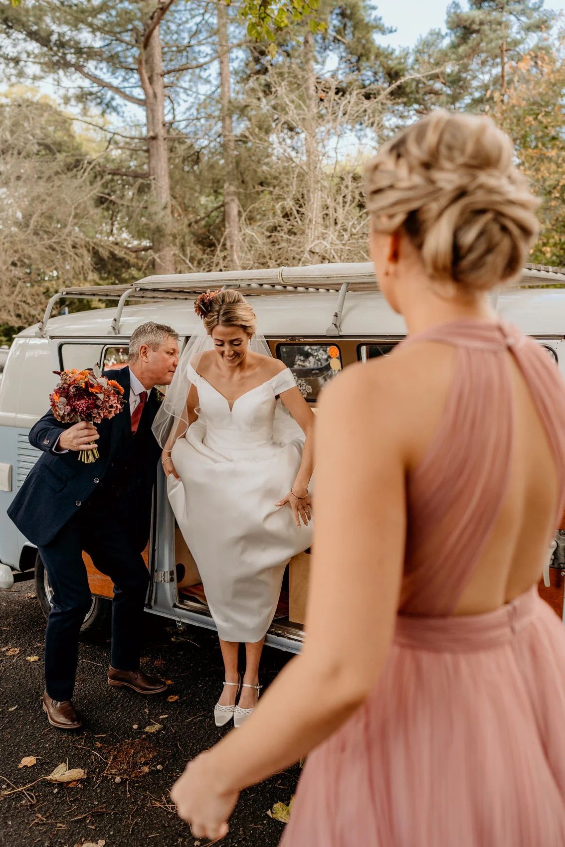 Wedding party gathered around a vintage car outdoors surrounded by trees and natural lighting.